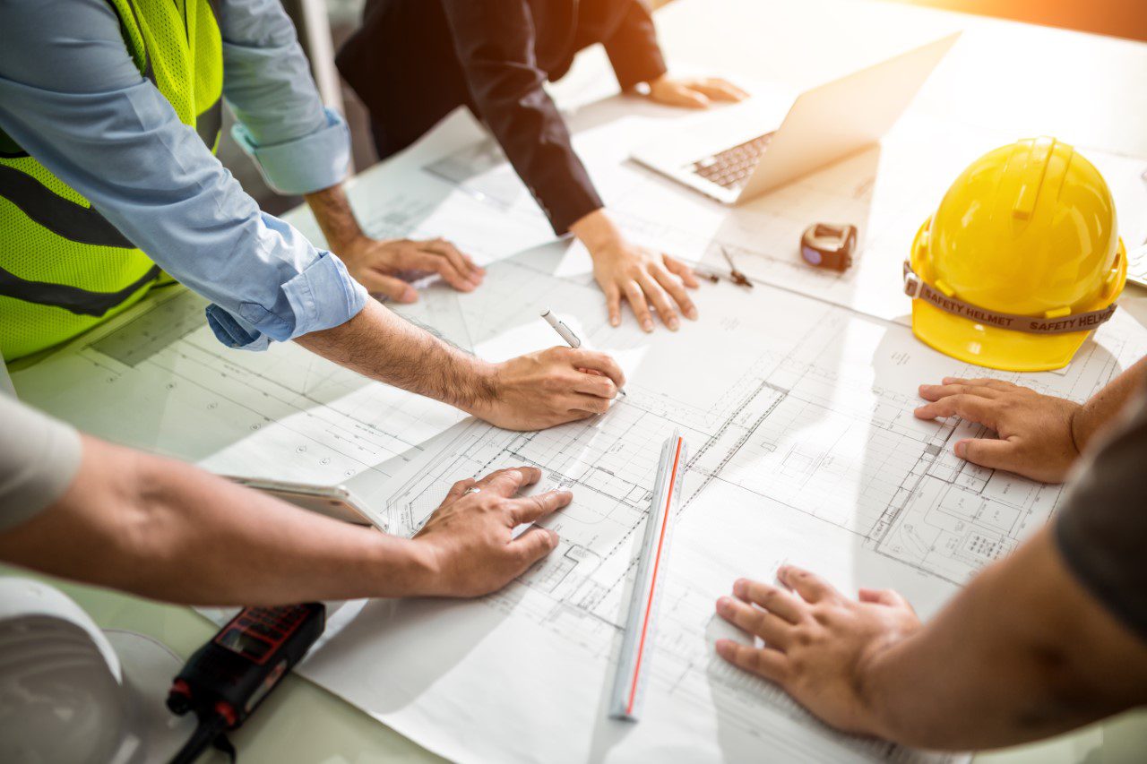 Several people collaborate on architectural blueprints at a table. A yellow safety helmet, rulers, and a laptop are visible.