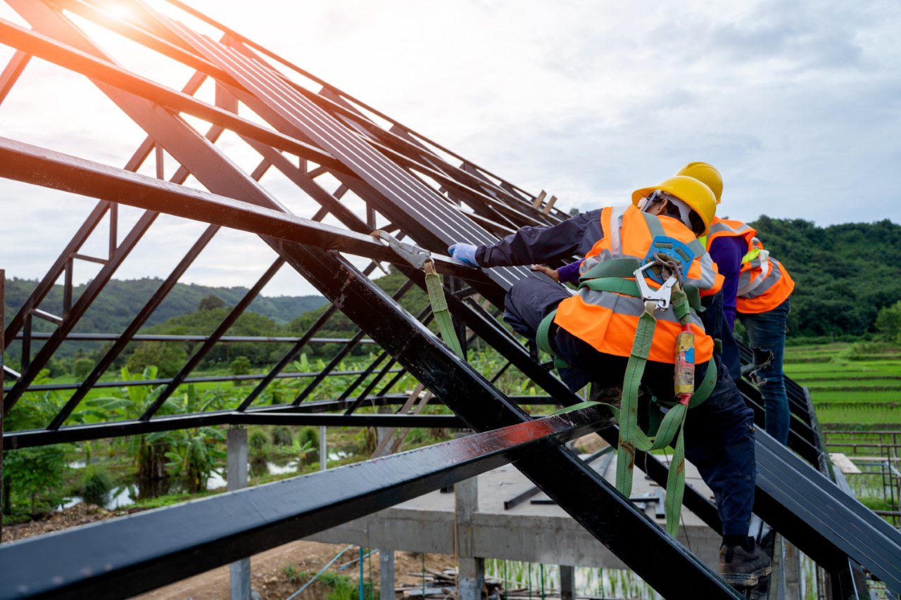 Two construction workers in safety gear are climbing a steel roof structure outdoors, with green fields and trees in the background.