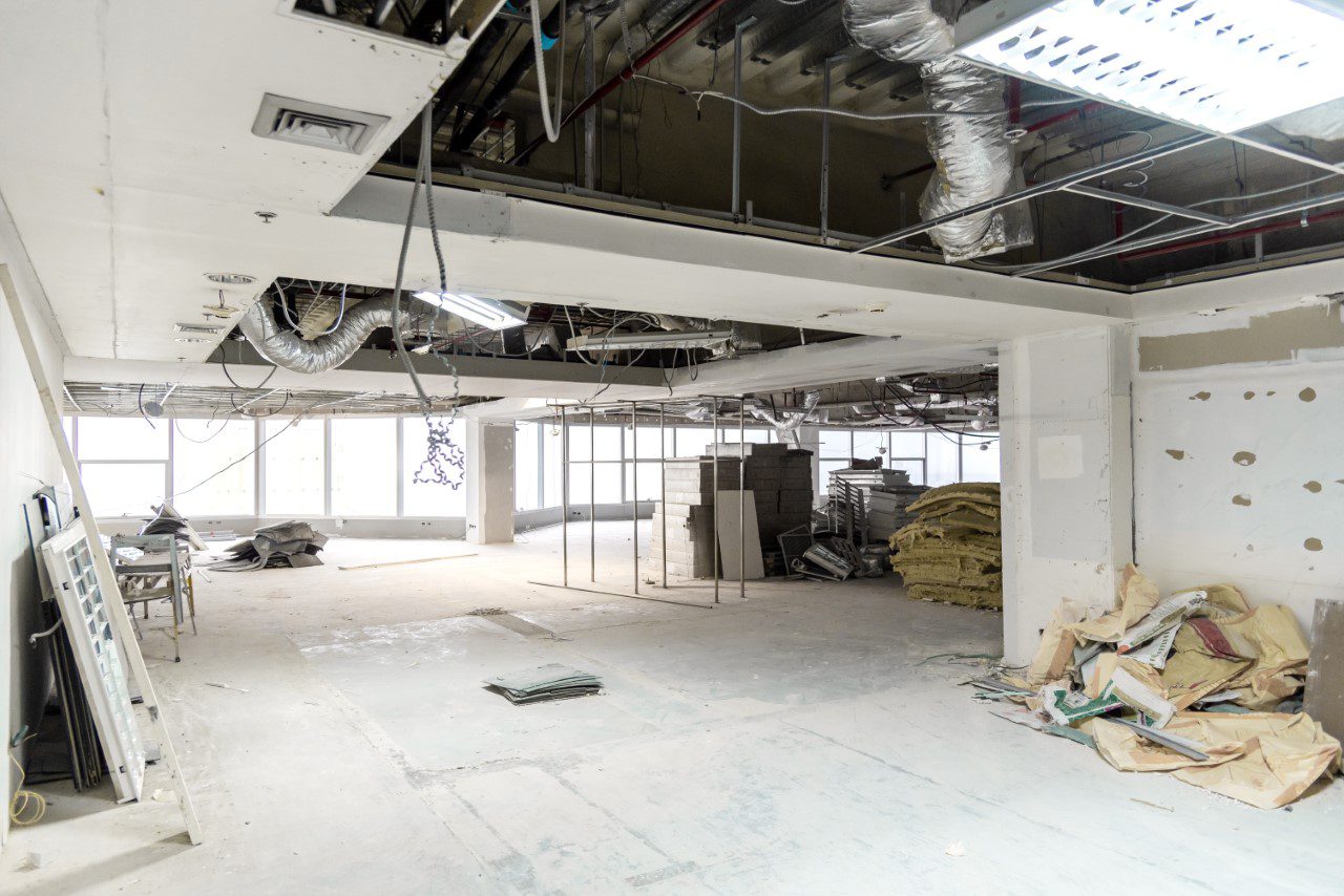 Unfinished room under construction with exposed ceiling wires, scattered building materials, and insulation stacked against walls. Large windows allow natural light to enter.